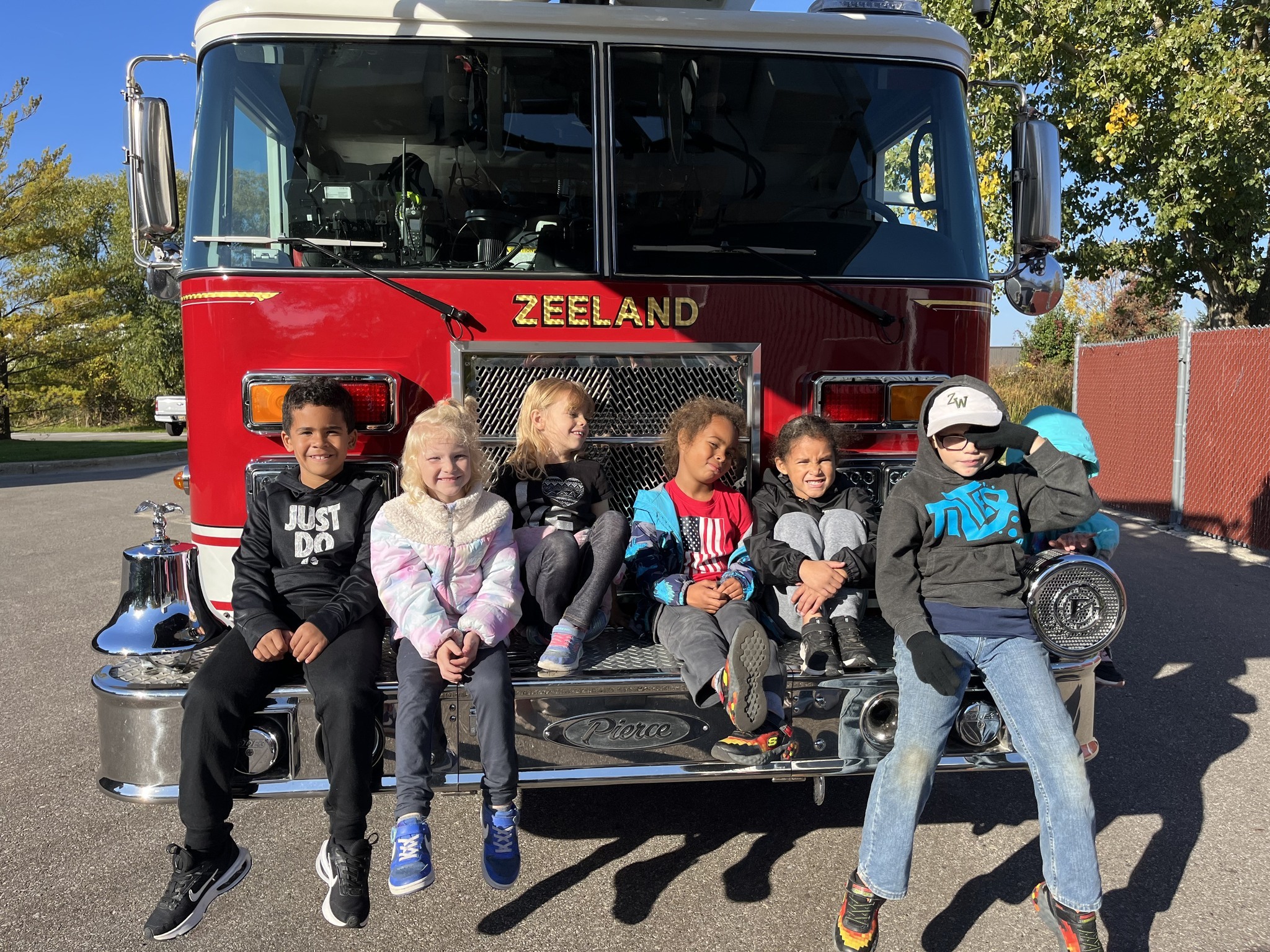 First grade students sit on the front bumper of a fire truck.